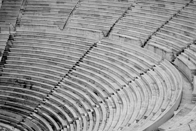 A photograph of seating rows in an ancient amphitheater. The stone rows and steps take up the whole frame leading from the lower right to the upper left in a curve.