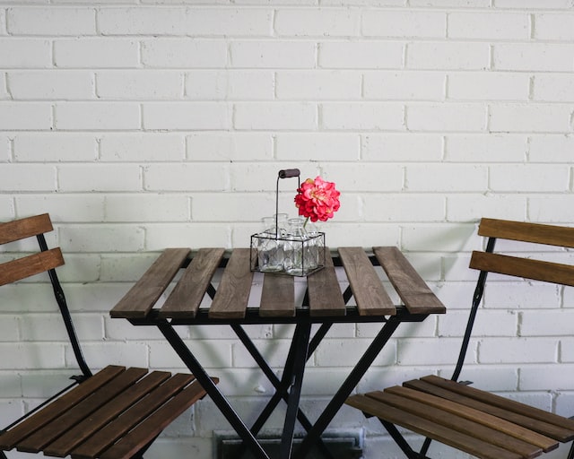 An image of a small wooden table with a flower on it. To the left and right are empty chairs. The scene is set in front of a white painted brick wall, which fills the whole background.